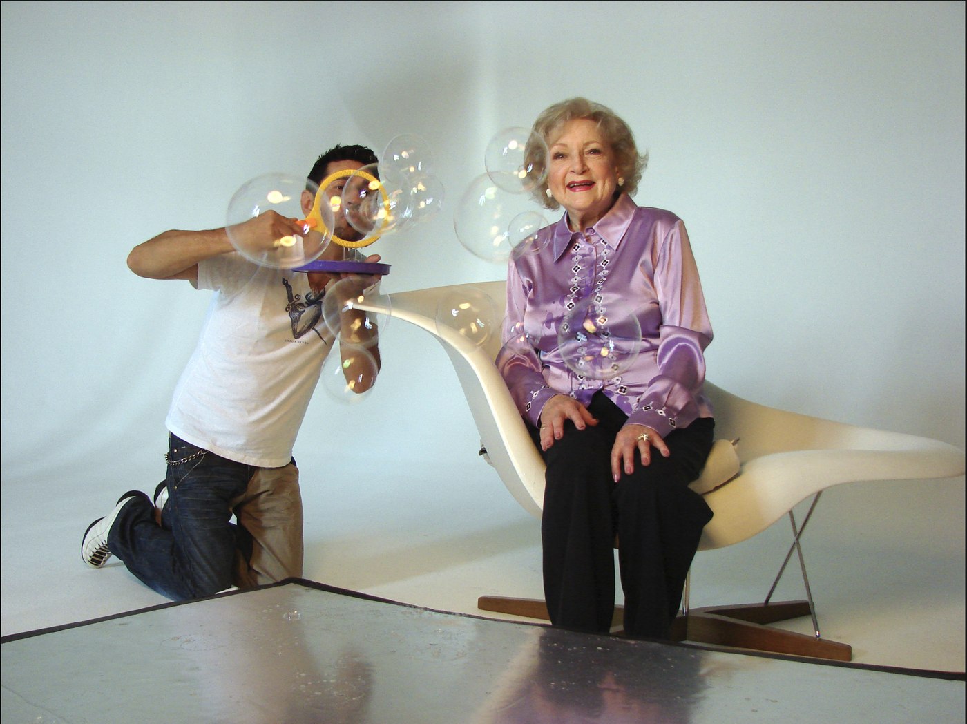 Behind the scenes on 'Life is Easy. Blow Bubbles.' — a crew member kneels to blow bubbles toward Betty White, seated in the white chair in the lilac silk blouse